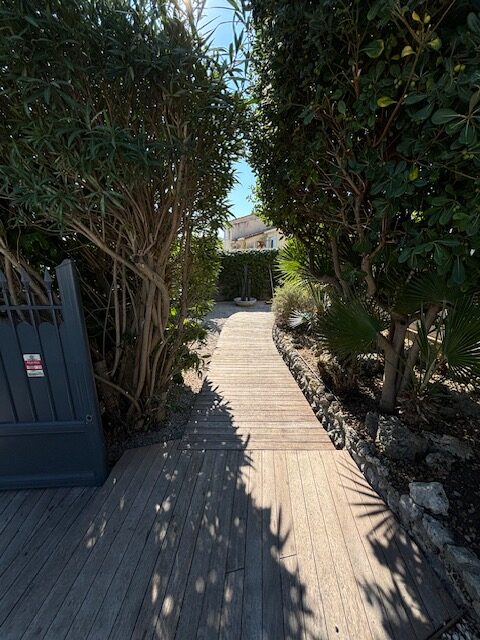 Allée en bois menant à une maison, bordée d'arbustes touffus et d'un portail gris à gauche avec une alarme rouge visible.