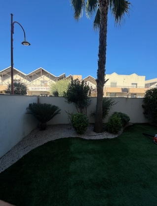 Jardin avec pelouse synthétique, palmier, arbustes et gravier blanc, devant un mur clair et des bâtiments dont un affiche "Le Jardin de la Raison".