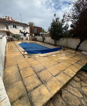Terrasse de piscine dallée de dalles jaunes et usées, avec une piscine recouverte d'une bâche bleue, près d'une maison et entourée de verdure.