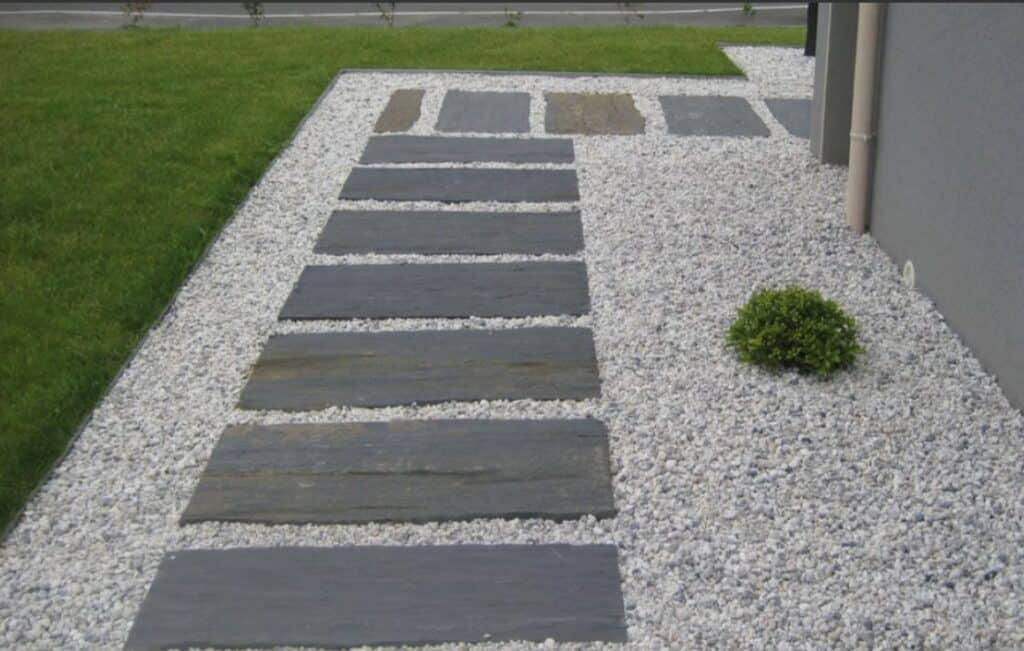 Allée Gravier Blanc et Pas Japonais Modernes Allée extérieure moderne avec pas japonais rectangulaires gris foncé sur gravier blanc, bordée de pelouse verte et d'un mur gris.