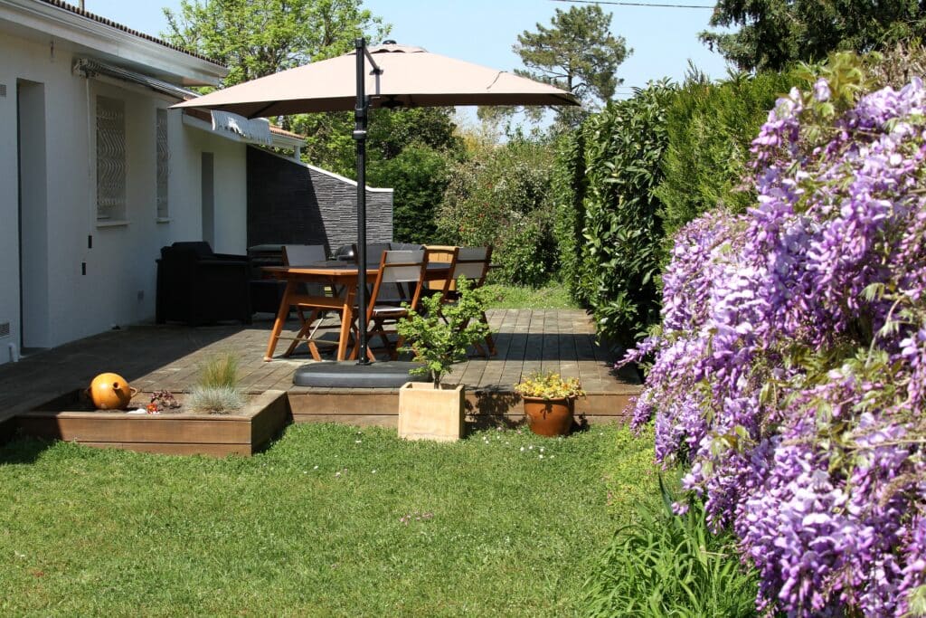Jardin fleuri et terrasse en bois ensoleillée Terrasse en bois aménagée avec table et chaises sous un parasol, près d'une maison blanche, entourée d'un jardin fleuri de glycine mauve.